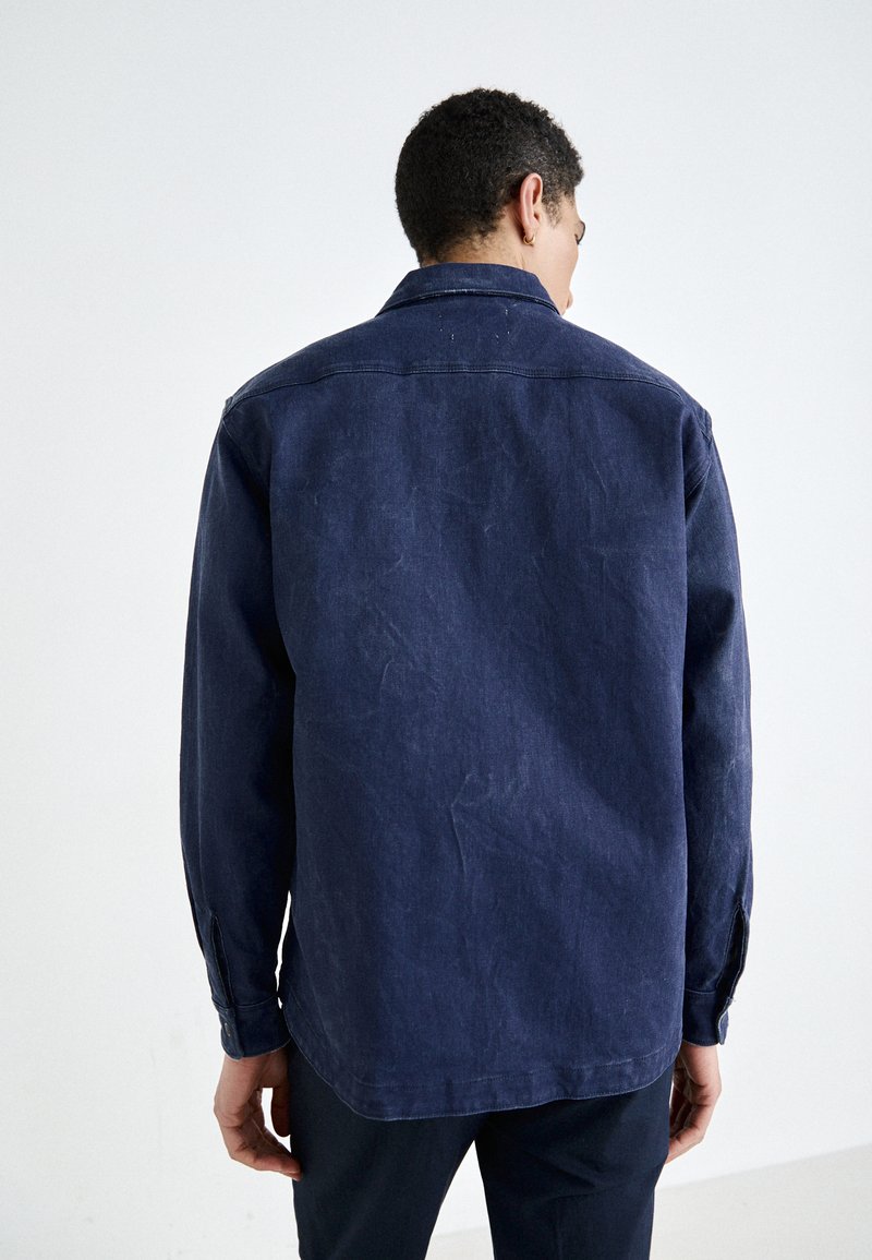 Granatowa koszula dżinsowa o luźnym kroju, z długimi rękawami, zapięciem na guziki przy mankietach oraz ze spiczastym kołnierzykiem. Delikatna faktura i lekko zużyty wygląd.