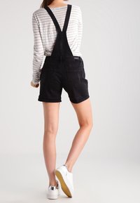Black denim overalls with a bib and adjustable straps, featuring a back pocket and cuffed shorts. Worn over a striped long-sleeve top.
