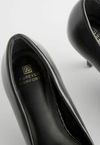 Czarne skórzane sandały na wysokim obcasie z zaokrąglonym noskiem, o gładkiej powierzchni, subtelnym przeszyciu oraz logo "Forever Comfort" w środku.