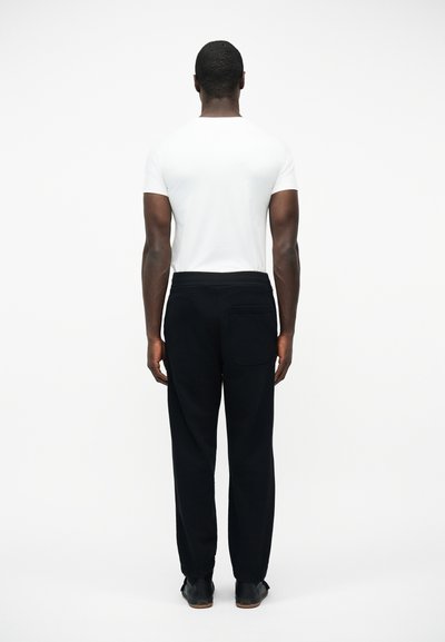 Homme debout, de dos, portant une chemise blanche à manches courtes, un pantalon noir et des chaussures noires, sur un fond blanc uni.