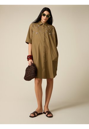 Kvinde iført olivengrøn kjole med knapper og blomsterbroderi, grønne solbriller, røde armbånd, holder en brun flettet taske og brune slidte sandaler.