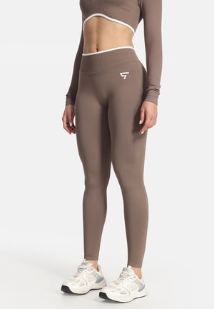 Donna che indossa leggings marroni a vita alta e crop top a manica lunga abbinato con rifiniture bianche e sneakers bianche da ginnastica.