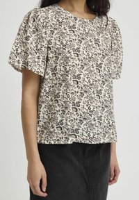 Bluse mit floralem Muster in Cremefarbe, kurzen Puffärmeln, rundem Ausschnitt und leichtem Stoff. Kombiniert mit einem dunklen Rock.