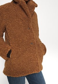 Brun fluffig kappa med hög krage, med en texturerad yta, två knappar och sidfickor. Bärs över blå jeans.