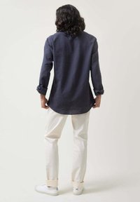 Camisa de lino azul marino con mangas largas, puños enrollados, dobladillo redondeado, combinada con pantalones crema y zapatillas blancas, vista desde la parte de atrás.