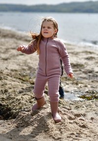 Lyserødt fleece-overdel med lynlås kombineret med brune gummistøvler, båret mens personen står på sandet strandterræn med vand i baggrunden.