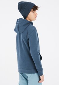 Felpa blu navy con cappuccio regolabile, tessuto morbido e tasca a marsupio, abbinata a un berretto a coste blu navy. Modello girato verso sinistra.