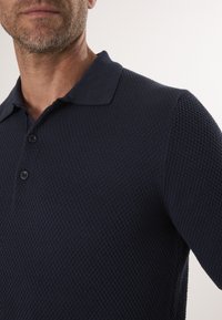Marine strikket polo med et struktureret mønster, tre knapper og flad krave. Tæt-fitting stil med en glat overflade og subtile detaljer.