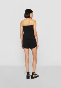 Sort, stropløs top med tekstureret elastisk bånd, parret med sorte shorts. Modellen har sorte stroppede sandaler på. Neutral baggrund.