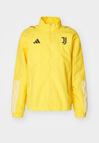 Chaqueta deportiva amarilla con cremallera frontal, cuello alto, acentos en blanco y negro, con tres franjas blancas en cada manga y un logo en el pecho.