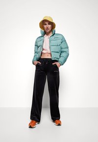 Lys blå pufferjakke, cropped rosa topp, svarte velour bukser og oransje sneakers. Modellen har på seg en gul bøttehatt, med hendene i lommene.