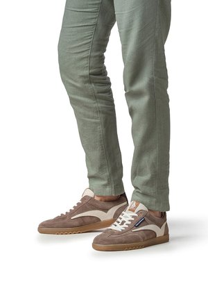 Persoon draagt olijfgroene broek en bruine suède sneakers met beige accenten en gumzolen, staand op een witte ondergrond.