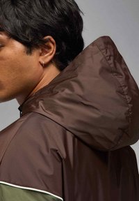 Ruskea hupullinen takki, jossa on sileä, kiiltävä pinta; siinä on pieni vihreä yksityiskohta ja valkea somiste alareunassa.