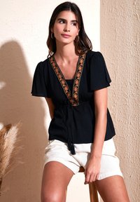 Zwarte blouse met korte pofmouwen, V-hals en bloemenborduursel langs de halslijn. Gepaard met lichte shorts.