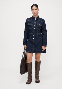 Γυναίκα που φοράει ένα σκούρο τζιν φορεματάκι με κουμπιά, κρατώντας μια μεγάλη καφέ δερμάτινη τσάντα, με καφέ μπότες μέχρι το γόνατο και χρυσά σκουλαρίκια κρίκους.