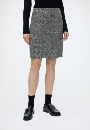 Frau trägt ein schwarzes langärmeliges Oberteil, einen knielangen schwarz-weißen gemusterten Rock, schwarze Socken und schwarze Lederschnallenschuhe.