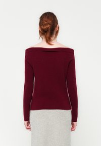 Γυναίκα με κόκκινα μαλλιά σε κότσο, ντυμένη με μπορντό εκρού πουλόβερ και ανοιχτό γκρι φούστα, στέκεται μπροστά σε λευκό φόντο.