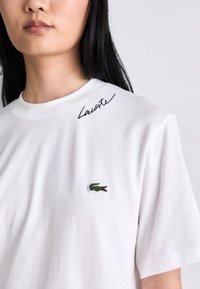 Žena v bílém tričku Lacoste s černým kurzívním logem poblíž límce a zeleným krokodýlem na hrudi.