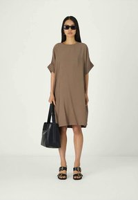 Mulher vestindo um vestido solto castanho, óculos de sol pretos, sandálias pretas, segurando uma bolsa preta, de pé contra um fundo claro liso.