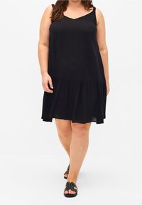 Žena jde v černých šatech bez rukávů s volánkem a v černých sandálech s otevřenou špičkou a stříbrnými nýty.