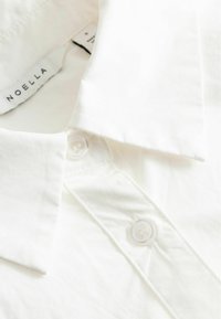 Gros plan d’un col de chemise blanc et d’une patte de boutonnage avec deux boutons transparents et une étiquette de marque Noella visible à l’intérieur du col.
