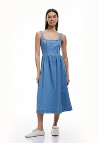 Denim midi-jurk met een vierkante halslijn, brede schouderbanden, geplooide taille en een uitlopende rok, gecombineerd met witte sneakers.