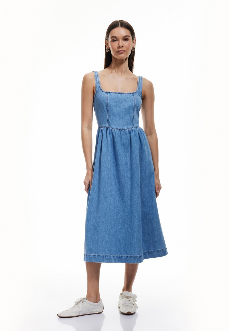Denim midi-jurk met een vierkante halslijn, brede schouderbanden, geplooide taille en een uitlopende rok, gecombineerd met witte sneakers.