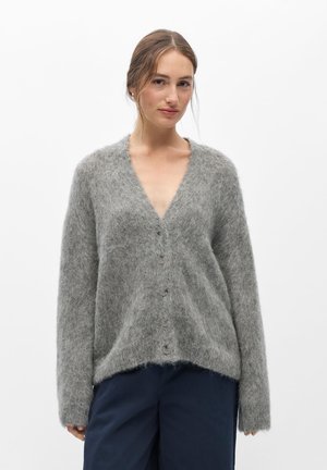 Femme aux cheveux châtain clair portant un cardigan gris à boutons, ample et duveteux, sur un pantalon bleu foncé, sur fond blanc uni.