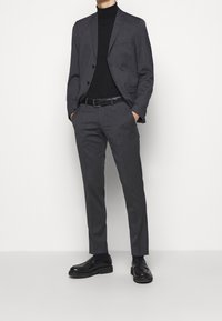 Abito grigio su misura con vestibilità slim, giacca monopetto e pantaloni abbinati. Indossato sopra un dolcevita nero, abbinato a scarpe in pelle nere.
