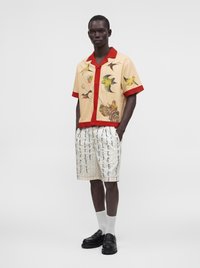 Homme portant une chemise beige avec bordure rouge et imprimés d’oiseaux, associée à un short blanc avec texte manuscrit bleu, chaussettes blanches et mocassins noirs.