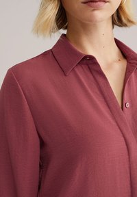 Camisa de botones color granate con cuello con solapas, confeccionada en tela texturizada. La camisa tiene un ajuste holgado y un detalle de un solo botón.