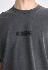 Camiseta de algodón gris con escote redondo. Presenta un logo impreso en negro en el lado izquierdo. La tela tiene una textura ligeramente desgastada.