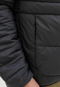 Jachetă puffer neagră, cu țesătură matlasată orizontal, având un buzunar lateral cu fermoar și detalii cu manșete ribbate. Textură interioară netedă.