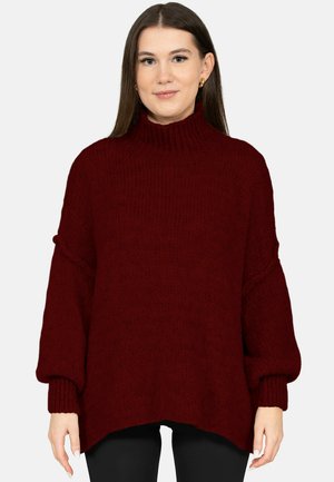 Kobieta z długimi brązowymi włosami, nosząca złote kolczyki koła oraz luźny, ciemnoczerwony sweter z golfem i czarne spodnie.