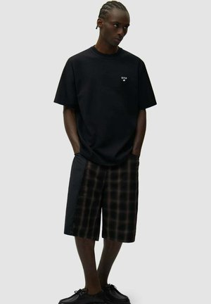 Uomo con capelli intrecciati che indossa una maglietta nera oversize e pantaloncini neri a quadri, con le mani nelle tasche, in piedi davanti a uno sfondo grigio uniforme.