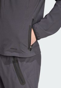 Grijze jas met een gestructureerde afwerking, voorzien van zwarte ritszakken en een elastische manchette, gelabeld "AEROREADY" op de zijkant.