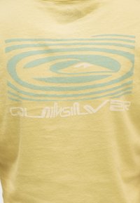 Κίτρινο ύφασμα με μπλε και λευκό στυλιζαρισμένο σχέδιο κύματος και κείμενο που μοιάζει με «Quiksilver» τυπωμένο στο κέντρο.