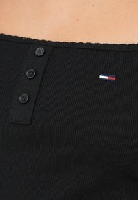 Maiou negru cu dungi, cu decolteu ondulat, trei nasturi negri și un mic logo brodat în roșu, alb și albastru.