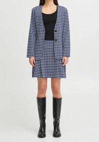 Femme portant un blazer et une jupe à carreaux bleus et blancs, avec un haut noir et des bottes noires montant jusqu'aux genoux, debout sur un fond uni.