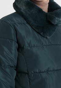 Teal fuskpälsjacka med en lyxig, mörk pälskrage, quiltat mönster och slät textur, med horisontella sömmar över kroppen.