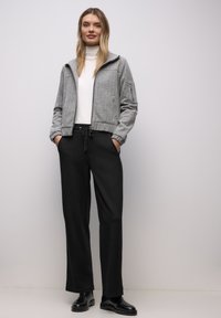 Chaqueta texturizada gris con cremallera y dos bolsillos, combinada con pantalones negros con cordón y botines negros, sobre un fondo neutral.