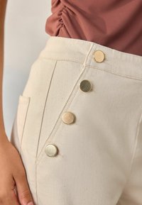 Witte katoenen broek met een zijrij van vier goudkleurige knopen. De stof heeft een gestructureerde afwerking en bevat een zijzak.