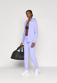 Ανοιχτό μωβ φούτερ με φερμουάρ και τσέπες καγκουρό, ταιριαστός παντελόνα φόρμας και μαύρο αθλητικό σουτιέν. Άσπρα αθλητικά παπούτσια και μαύρη τσάντα duffel.