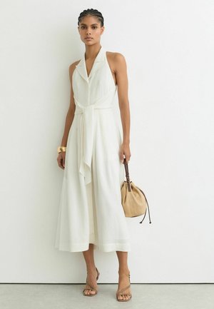 Naine punutud juustega, kandes valget varrukateta midi-kleiti, hoides käes beežikat käekotti ning kandes kuldset käevõru ja rihmikuid.