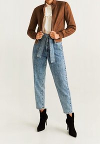 Veste bomber en daim marron, blouse blanche à volants, jeans taille haute bleu clair avec ceinture, et bottines noires à talons aiguilles.