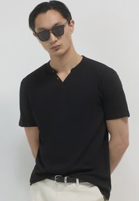 Maglietta nera a maniche corte con scollo a V e dettaglio con bottoni, abbinata a occhiali da sole scuri. Il modello si trova con le mani dietro la schiena su uno sfondo neutro.