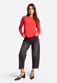 Rotes Langarmshirt mit abgerundetem Saum, kombiniert mit hochgeschnittenen, dunkelblauen, cropped Jeans. Accessoires sind schwarze Riemchenschuhe.