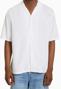 Man i en enkel vit kortärmad skjorta med knappar och en camp collar, samt ljusblå denimjeans, står mot en vit bakgrund.