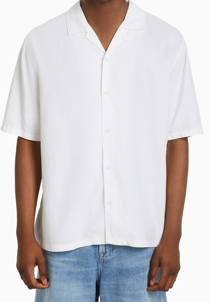 Man i en enkel vit kortärmad skjorta med knappar och en camp collar, samt ljusblå denimjeans, står mot en vit bakgrund.