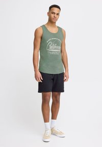 Zeleni top bez rukava s otisnutim logotipom "California", uparen s crnim kratkim hlačama, bijelim čarapama i svijetlosmešavim tenisicama; ležeran atletski dizajn.
